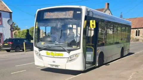 A white X91 "Chew Valley Sprint" bus with the words "The Big Lemon" on the front, waiting at a bus stop with its doors open. A residential street can be seen in the background.