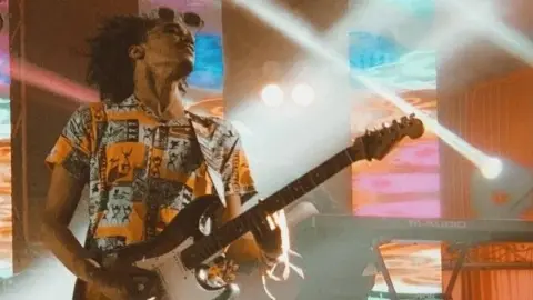 A man wearing a black, white and orange patterned shirt playing a guitar on stage with strobe lights shining behind him.