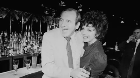 Getty Images A black and white image of actor Leonard Rossiter and Joan Collins laughing together at a bar. He is on the left and wearing a light-coloured blazer, shirt and tie. She is on the right and has short curling dark hair and wearing a dark blouse
