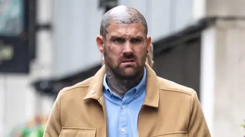 PA Media Tony Bartlett walks towards Bristol Crown Court. He is a large man with a shaved head and several tattoos on his neck and head, but not on his face itself. He is wearing a light blue shirt under a light brown jacket