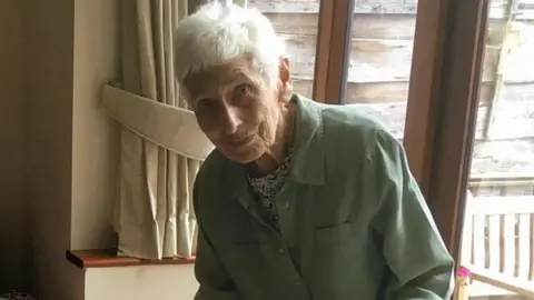 Family photograph Judy Zussman, with short grey hair and wearing a black and white top with a mint green coat, is photographed standing in front of a window with cream tied-back curtains.