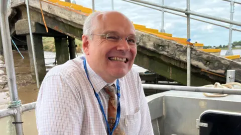 Michael de Whalley sitting on a small boat. He is wearing a white, collared shirt which has a checked pattern of thin multi-coloured lines. He is also wearing a patterned tie and a blue lanyard round his neck. He is looking directly at the camera, is smiling and is wearing a pair of glasses. There is a stepped gangway behind him leading down to the boat he is in. The tide is out under the gangway revealing the muddy shore.