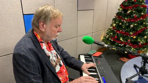 Rick Wakeman sitting at his piano in the BBC Radio Suffolk studio. There is a microphone in front of him. A Christmas tree is in the background. 