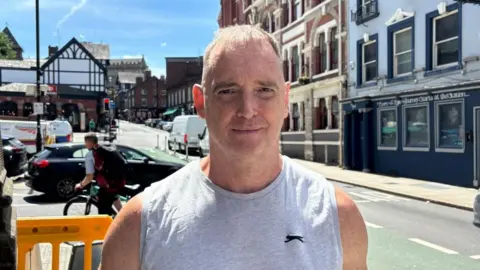 Charlie Stubbs/BBC There is a man in gym clothes. He has a grey sleeveless Slazenger shirt on. He has thin light hair. Behind is the gyratory system which includes a car which has stopped inside of the green cycle path.