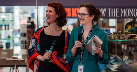 Royal Armouries Two women standing in a museum with microphones, smiling. One wears a black top and red shawl and the other a green dress. 