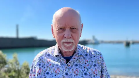 John Baker is staring into the camera while stood in front of Elizabeth Harbour. The sea and Albert Pier are out of focus behind him. He is wearing a white floral shirt.