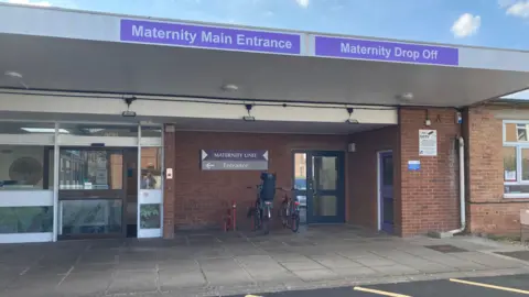 The outside of a single storey brick building. There are three doors into the building, with two bikes outside. Above the building are two purple signs saying 'maternity main entrance' and 'maternity drop off'.