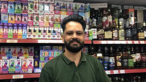 ALEX STEVANOVIC/BBC Parmjit is standing in his shop with various products on sale behind him.