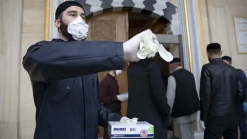 Getty Images Volunteers in face masks hand out gloves at the entrance to the Heart of Chechnya Mosque in Grozny, Chechen Republic