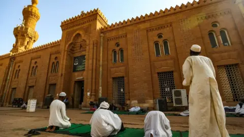 Getty Images A man dressed in white gazing up at the mosque. The mosque is a sandy colour, with intricate designs and there are blue skies.