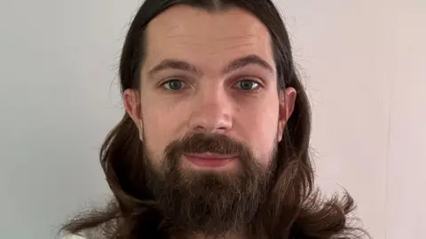 Alex Cole Alex Cole with a full beard and long hair looking at the camera