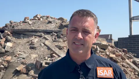 Lang stands in front of a pile of bricks, concrete and rubble He wears a dark top with UK Fire and Rescue ISAR orange badge.