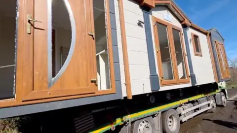 The mobile home in its unspoiled form on the back of a trailer. It has white walls with brown doors, windows and eaves.