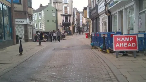 A dry Saddler Street after pipe and road repairs. A patch of the road where a water pipe burst has been filled with asphalt concrete. Small rectangle stones cover the rest of the road. Part of the right side of the street is fenced off with a read sign reading Ramp Ahead. A small group of people is walking down the street.
