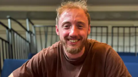 A blond man wearing a brown t-shirt. He is looking at the camera and smiling.