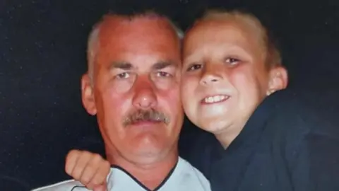 Callum Dickson A young boy with short brown hair and wearing a dark-coloured T-shirt with a red, white and blue design on the front is smiling while his arm is around his father's shoulder. HIs father has short brown hair and a moustache and is wearing a white football shirt.