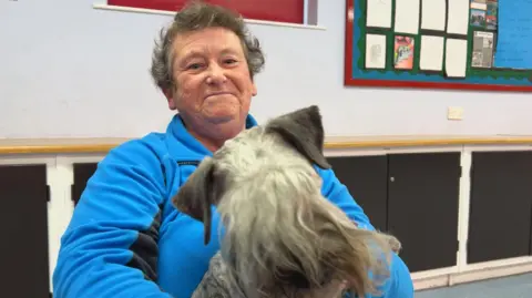 Christine is wearing a bright blue fleece sits indoors holding a small, grey, shaggy-haired dog. The room has light-colored walls, a display board with papers on the right, and dark cabinets along the back wall.