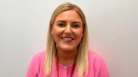Kathryn Dobbin has long blonde hair and is wearing a pink cardigan. She is standing in front of a plain white wall. 
