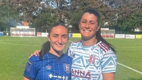 Sophie Peskett and Holly Turner, in opposing coloured kits, arm in arm after their most recent league clash by the side-lines. 