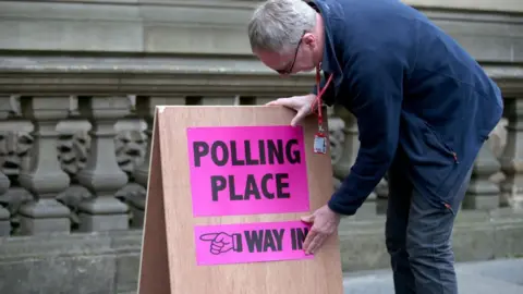 A man with grey hair, wearing dark clothes, bends over a sign that says "polling place". The sign is on the pavement of a street, with a stone railing in the background. 