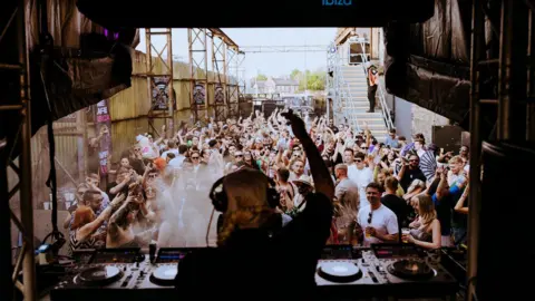 KoLAB Studios A female DJ stands behind some decks with one hand in the air and headphones on her head. She performs to a crowd of people outside of a warehouse