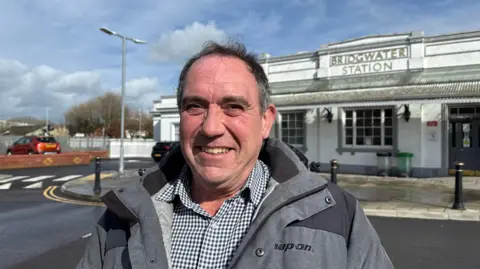 Man with dark grey hair looking at the camera. He's stood in front of the Bridgwater Railway Station sign which is an old Victorian white building.