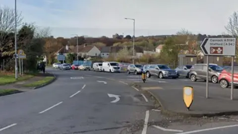 A wide entrance to a road off a roundabout, with a queue of traffic on the other side of the road. A sign for Cannock and Stafford can be seen on the right