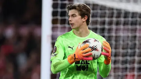 Melker Ellborg holding a ball in two hands close to his chest. He has dark hair and is wearing a lime green Malmo shirt and orange Puma goalkeeping gloves. His goal can be seen behind him.