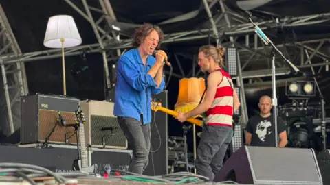 Razorlight on stage. Singer Johnny Borrell is in a blue shirt and dark jeans, and is singing intensely into the microphone with his eyes screwed shut.