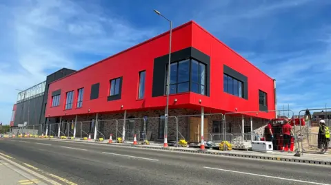Tom MacDougall/BBC The exterior of the building, which has bright red panelling and large windows, surrounded by construction fencing.