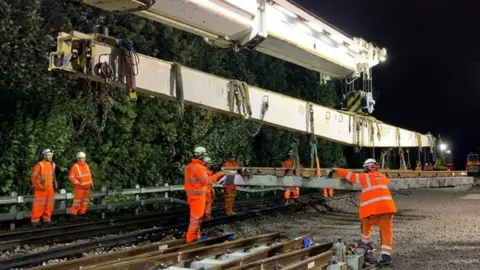 Network Rail Rail engineering workers in orange hi-vis suits and white hard hats guiding a steel girder hanging from a crane