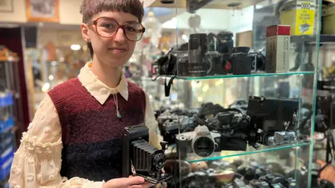 Oliver holds the antique camera and smiles in front of Salisbury Antique Market's wide collection of old photography items.