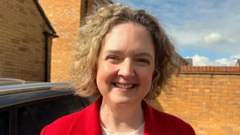 The picture shows Anna Sabine smiling into the camera. She is wearing a red jacket. There is a brick wall behind her and a car to the left of the picture.
