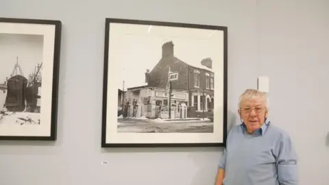 Alec Gill An elderly man in glasses and a light blue sweater standing beside a framed black and white photograph in a gallery. The photo shows an old Rix petrol station with pumps and brick buildings.