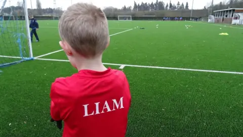 Chris Priday Liam facing away from the camera looking out at a pitch. On the back of his red shirt is his name in capital letters, akin to the names on the back of footballers' shirts.