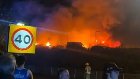 A grass fire is seen in the distance as people watch by a street.