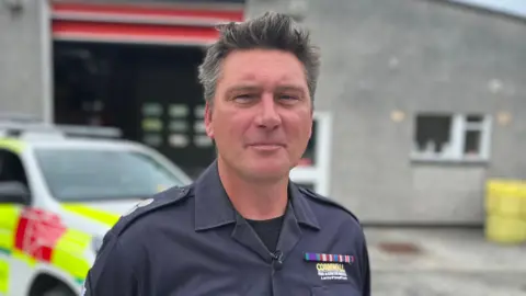 A man in fire uniform looks straight to camera, he is standing in front of a fire station and co-responding 4x4 type vehicle.