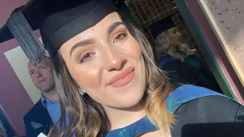 Sophie Morgan wears a black university cap and gown and smiles at the camera. She has brown short hair and brown eyes. 