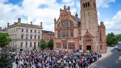 Aodhán Roberts A crowd of people gathered in front of Derry's Guildhall for an anti-racism rally. There is blue sky.