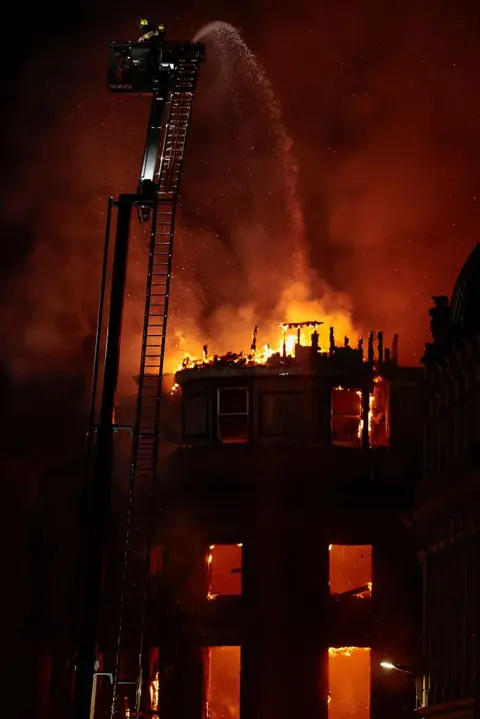 Getty Images A burning and destroyed building, with firefighters on an extendable ladder spraying water over it
