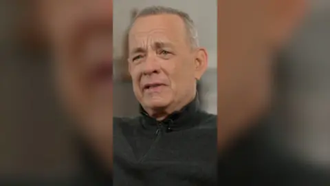 Tom Hanks, close up, wearing a black sweater. He's mid speech.