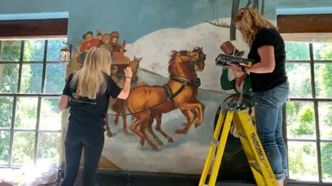 BBC Artists Sarah Daniels and Sarah Hodgkins restore part of the mural showing a coach struggling up the road from Scotch Corner in the snow
