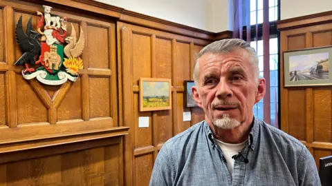 Councillor Richard Brown, Labour's Cabinet Member for Finance, stands in front of the council coat of arms. He is in a wood panelled room and wears an unbuttoned blue denim shirt with a white t shirt underneath.