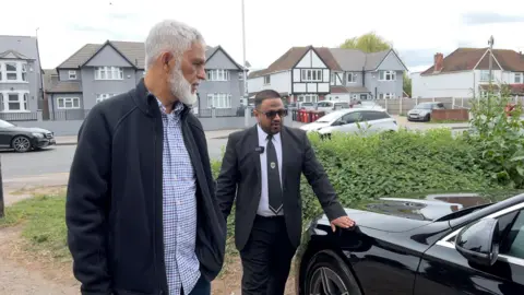 Two men, one in casual clothes with a trimmed grey beard, the other in a  chauffeur's uniform, inspect a black Mercedes car.