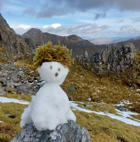 Liam Turbett A melting snowman sitting on a rock,  on green hills 