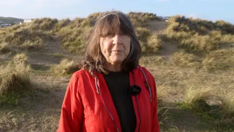 A woman with dark shoulder length hair wearing a red rain jacket, with sand dunes behind her
