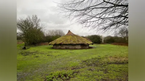A roundhouse in a field surrounded by trees and shrubs.