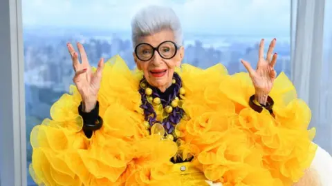American fashion designer Iris Apfel. She is seen wearing a puffy yellow dress and holding both hands in the air.