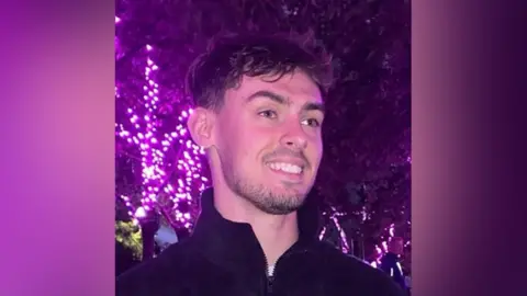 Sam Eklls A man with short, dark tousled hair wears a high-necked zip-up fleece as he stands, smiling, in front of a tree decorated with bright purple lights.