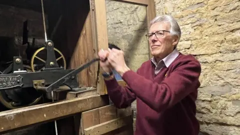 Kate Bradbrook/BBC Man wearing red sweater pulls on the handle of a clock mechanism inside a stone tower